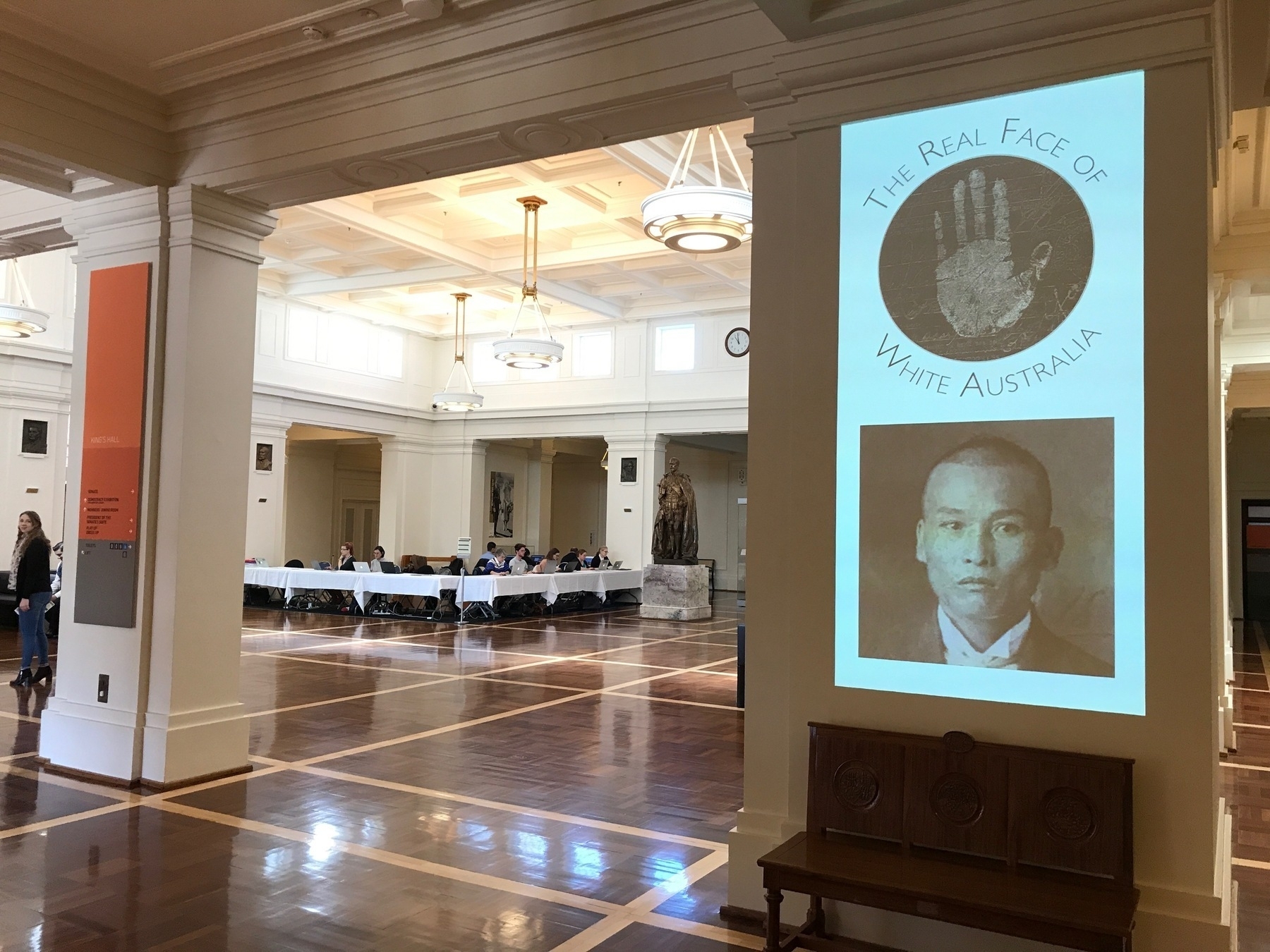 Photograph of Kings Hall in Old Parliament House. In the foreground, the Real Face of White Australia logo, and a portrait photo from one of the records, is projected on a column. In the background are tables where volunteers are busily transcribing records.